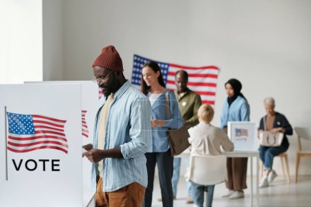 Photo for Group of young multicultural voters in casualwear standing in queue along vote booths in polling place and putting their ballots into boxes - Royalty Free Image