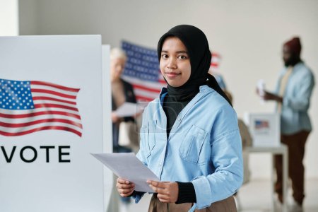 Téléchargez les photos : Jeune femme musulmane souriante dans le hijab tenant un bulletin de vote et regardant la caméra tout en se tenant contre les électeurs dans un bureau de vote - en image libre de droit