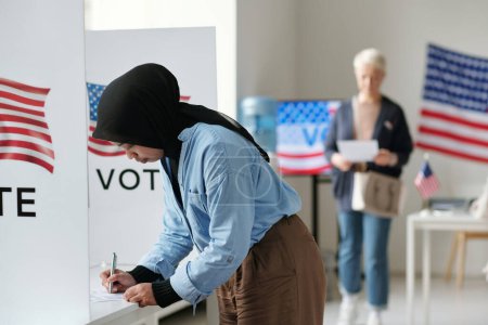 Foto de Mujer musulmana joven en hiyab inclinándose sobre la cabina de votación y haciendo su elección de candidato durante las elecciones presidenciales en el lugar de votación - Imagen libre de derechos