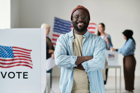 Téléchargez les photos : Heureux jeune homme afro-américain en tenue décontractée regardant la caméra avec le sourire tout en se tenant au bureau de vote contre les électeurs interculturels - en image libre de droit