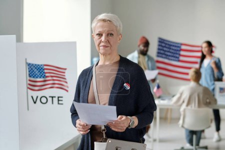 Photo for Confident mature blond female voter with ballot paper looking at camera while standing in polling place full of American citizens - Royalty Free Image
