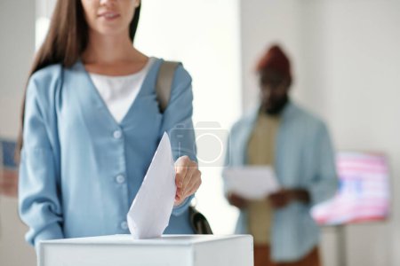 Photo for Focus on hand of young female voter putting ballot paper into box while standing in front of camera against black man in polling place - Royalty Free Image
