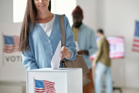 Foto de Foto recortada de mujer joven en ropa de casualwear fundiendo papeleta de votación en una caja blanca con bandera estadounidense mientras está de pie delante de la cámara - Imagen libre de derechos