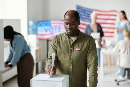 Photo for Mature African American man taking part in presidential elections while standing by ballot box and putting document with his choice there - Royalty Free Image