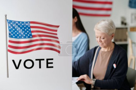 Photo for Part of voting booth with American flag and two female voters sitting behind and choosing candidate from list on ballot papers - Royalty Free Image