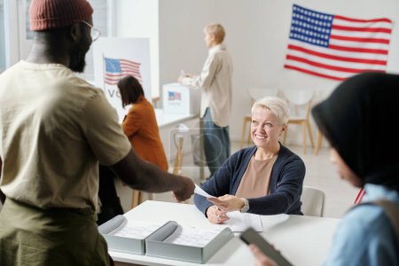 Téléchargez les photos : Joyeux blond femme mûre passer le bulletin de vote au jeune homme afro-américain debout par bureau de la commission électorale - en image libre de droit