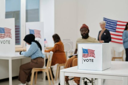 Téléchargez les photos : Focus sélectif sur bureau avec urne pour les bulletins de vote avec des candidats choisis par les électeurs sur fond de personnes interculturelles faisant leur choix - en image libre de droit