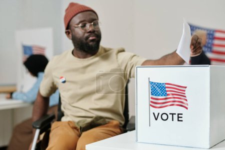 Téléchargez les photos : Boîte pour bulletins de vote sur bureau et jeune Afro-Américain handicapé assis en fauteuil roulant et faisant son choix - en image libre de droit
