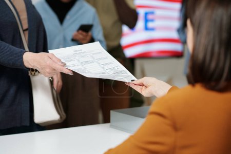 Téléchargez les photos : Gros plan d'une jeune femme de la commission électorale qui transmet un bulletin de vote à un électeur en file d'attente devant le bureau d'inscription - en image libre de droit