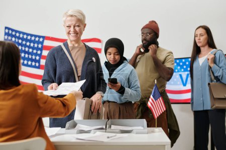 Foto de Cola de votantes interculturales de pie junto a la mesa de la comisión electoral mientras uno de ellos toma papeletas con la lista de candidatos - Imagen libre de derechos