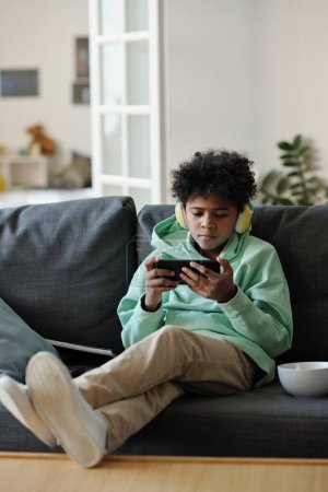 Photo for African American schoolboy in casualawear watching online movie or video in smartphone while having rest on comfortable couch at home - Royalty Free Image