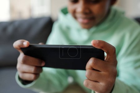 Téléchargez les photos : Focus sélectif sur les mains du petit garçon tenant son téléphone portable devant la caméra tout en regardant une vidéo ou un film en ligne à loisir - en image libre de droit