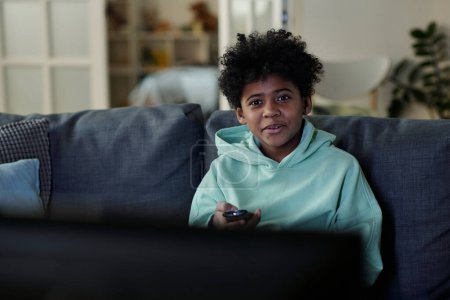 Téléchargez les photos : Jeune garçon assis sur le canapé devant le téléviseur et regardant à travers les canaux à l'aide de la télécommande tout en choisissant quelque chose de curieux - en image libre de droit