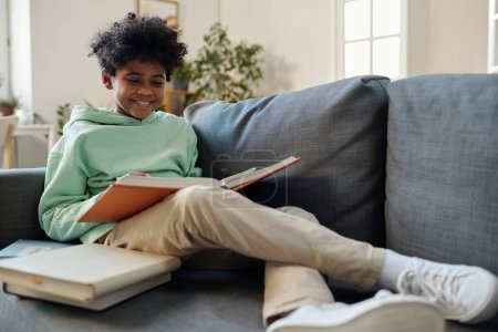 Téléchargez les photos : Adolescent écolier en causalvêtements assis sur le canapé et la lecture de l'encyclopédie tout en se préparant pour la leçon de géographie ou de biologie - en image libre de droit