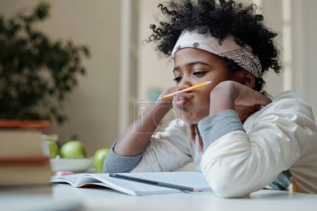 Téléchargez les photos : Écolier distrait et paresseux réticent à faire ses devoirs assis par bureau avec un copybook ouvert et tenant un crayon entre le nez et la lèvre supérieure - en image libre de droit
