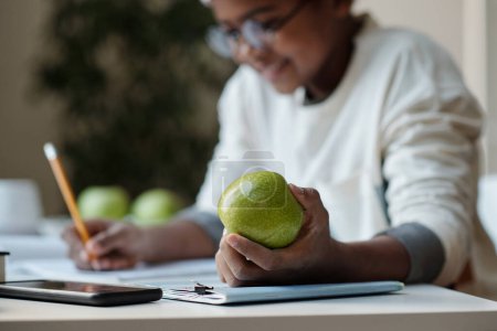 Photo for Focus on hand of diligent schoolboy holding fresh green apple while sitting by desk at home and carrying out home assignment - Royalty Free Image