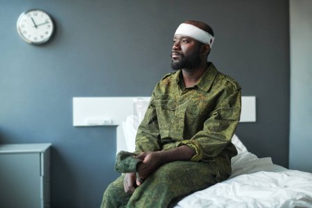 Photo for Young African American stressed soldier with bandaged head sitting on bed in hospital ward, looking through window and thinking - Royalty Free Image