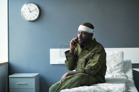 Photo for Young African American soldier in camouflage sitting on bed in hospital ward and talking to his friend or family on mobile phone - Royalty Free Image