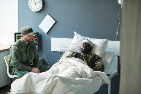 Foto de Joven en uniforme militar sentado frente a su amigo con la cabeza vendada y gotero acostado en la cama mientras lo visita en la sala del hospital - Imagen libre de derechos