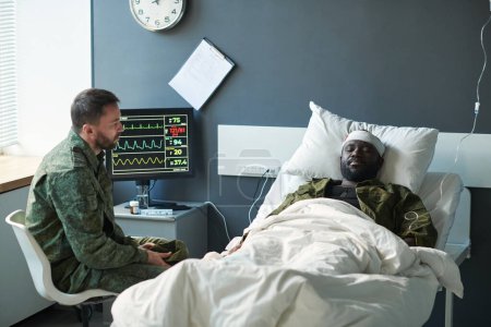 Foto de Soldado afroamericano herido con la cabeza vendada acostado en la cama en el hospital mientras su amigo en camuflaje sentado frente a él - Imagen libre de derechos
