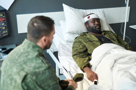 Foto de Enfoque del joven afroamericano herido en camuflaje acostado en la cama en las salas del hospital mientras su amigo se sienta frente a él durante la visita - Imagen libre de derechos
