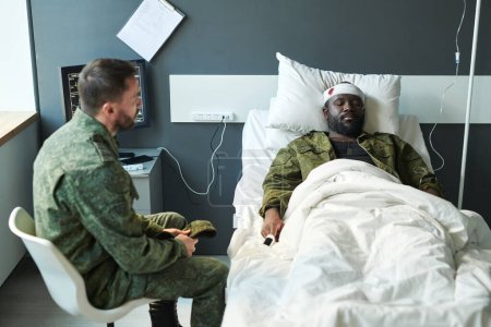 Foto de Joven soldado en uniforme militar sentado junto a la cama de su amigo en coma con gotero cerca mientras lo visita en el hospital - Imagen libre de derechos