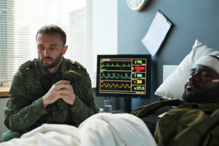 Foto de Joven soldado en uniforme militar sentado junto a la cama de un amigo afroamericano herido con la cabeza vendada mientras lo visitaba en el hospital - Imagen libre de derechos