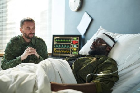 Foto de Joven soldado afroamericano con la cabeza vendada acostado en la cama de hospital mientras su amigo en uniforme militar sentado frente a él durante la visita - Imagen libre de derechos
