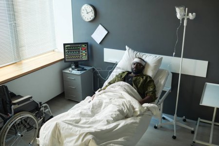 Foto de Joven afroamericano herido en camuflaje acostado en la cama entre el gotero y la pantalla de la computadora con indicadores médicos - Imagen libre de derechos