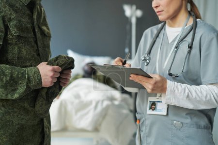 Photo for Close-up of young doctor or nurse in medical scrubs making prescriptions in document while standing in front of soldier visiting his sick friend - Royalty Free Image
