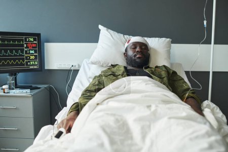 Foto de Soldado afroamericano con la cabeza envuelta por un vendaje acostado en la cama entre el gotero y la pantalla de la computadora con indicadores médicos - Imagen libre de derechos