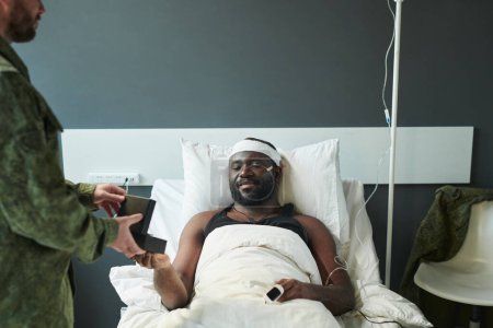 Foto de Joven herido acostado en la cama y mirando la medalla en una pequeña caja sostenida por un soldado en uniforme militar de pie frente a su amigo - Imagen libre de derechos