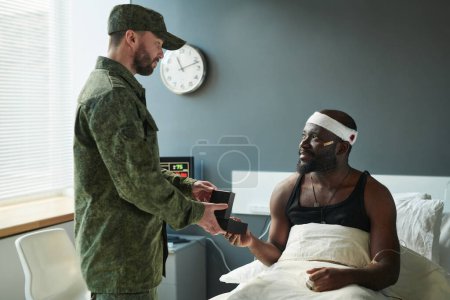 Photo for Young injured soldier with bandaged head sitting on bed in front of his friend passing him small box with medal during visit in hospital - Royalty Free Image