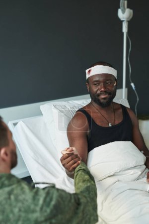 Foto de Joven soldado herido con la cabeza vendada sentado en la cama en la sala del hospital militar y mirando a su amigo durante el apretón de manos - Imagen libre de derechos