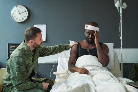 Photo for Young man in military uniform supporting his injured friend sitting on bed and keeping hand on wounded head wrapped with bandage - Royalty Free Image