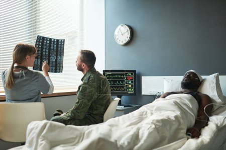Téléchargez les photos : Jeune médecin expliquant les détails du scanner cérébral du patient blessé au soldat en uniforme militaire alors que les deux assis devant la fenêtre dans la salle - en image libre de droit
