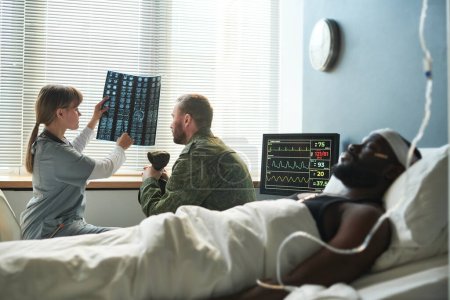 Foto de Enfermera en matorrales médicos explicando los resultados de rayos X a soldado en uniforme militar mientras está sentado en la sala del hospital con el paciente lesionado - Imagen libre de derechos