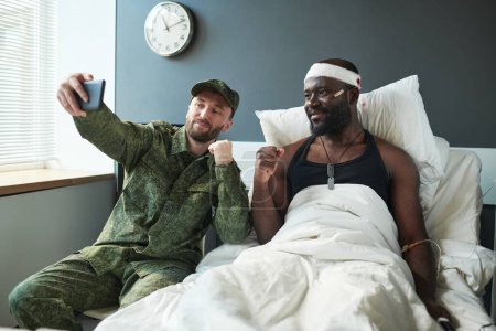 Foto de Dos jóvenes soldados interculturala tomando selfie en la sala del hospital mientras uno de ellos sentado junto a la cama de un amigo herido con la cabeza vendada - Imagen libre de derechos