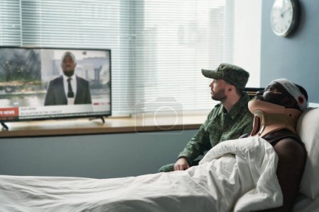 Téléchargez les photos : Vue latérale de l'homme blessé et son copain en uniforme militaire regardant les nouvelles diffusées à la télévision alors qu'ils étaient assis devant l'écran dans la salle d'hôpital - en image libre de droit