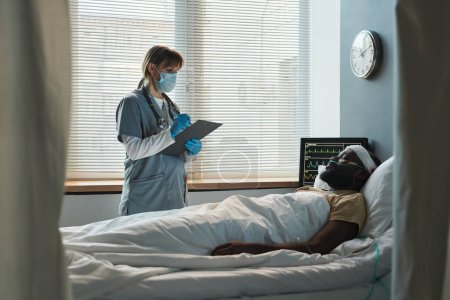 Téléchargez les photos : Jeune homme afro-américain blessé avec des bandages tête couchée au lit pendant que l'infirmière fait des ordonnances dans la salle d'hôpital - en image libre de droit