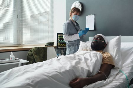 Photo for Young injured man with oxen nasal cannula lying in bed against nurse in medical scrubs and protective mask standing by computer - Royalty Free Image