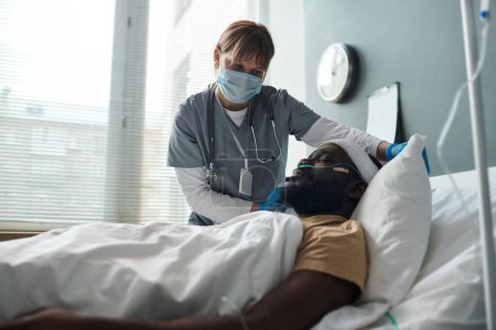 Téléchargez les photos : Infirmière en masque de protection redressant l'oreiller d'un patient afro-américain blessé avec la tête pansée couchée dans son lit dans un hôpital militaire - en image libre de droit