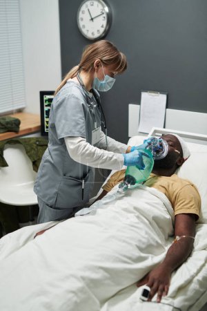 Foto de Médico en exfoliantes médicos cambiando globo de oxígeno y mascarilla chacking en ventilación pulmonar artificial mientras está de pie junto a la cama con el paciente - Imagen libre de derechos