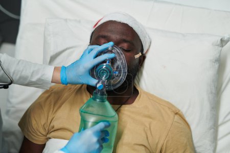 Photo for Gloved hand of nurse or caregiver holding oxygen mask and balloon helping sick male patient with bandaged head breathe - Royalty Free Image