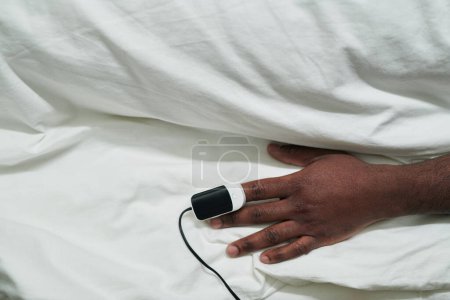 Foto de Mano de joven afroamericano paciente masculino con oxímetro de pulso en la punta del dedo acostado bajo sábana de algodón blanco en la cama en la sala de hospital - Imagen libre de derechos