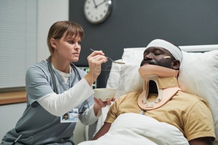 Foto de Enfermera en un tazón uniforme con gachas mientras alimenta a un paciente lesionado en cabeza vendada y cuello médico en el cuello en la sala de hospital - Imagen libre de derechos