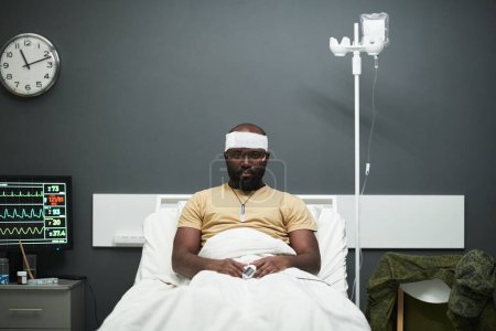 Foto de Joven soldado afroamericano con oxímetro en la punta de los dedos y vendaje en la cabeza sentado en la cama en la sala de hospital y mirando a la cámara - Imagen libre de derechos