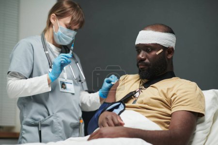 Photo for Young injured man with bandaged head and broken arm sitting in bed while nurse preparing his shoulder for injection in military hospital - Royalty Free Image