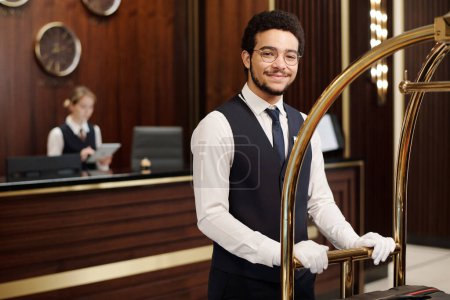 Photo for Young elegant bellboy in uniform pushing cart with clients baggage while moving along hotel lounge and looking at camera - Royalty Free Image