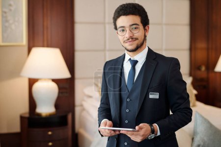 Photo for Young confident head manager of luxurious hotel looking at camera while standing against lamp and comfortable double bed in room - Royalty Free Image